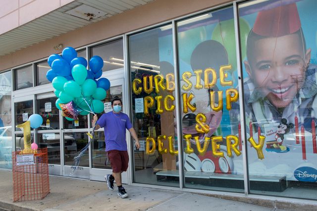 A man holds a bunch of balloons outside a PartY City store that says "Curbside Pickup Available" in the window (in balloons)